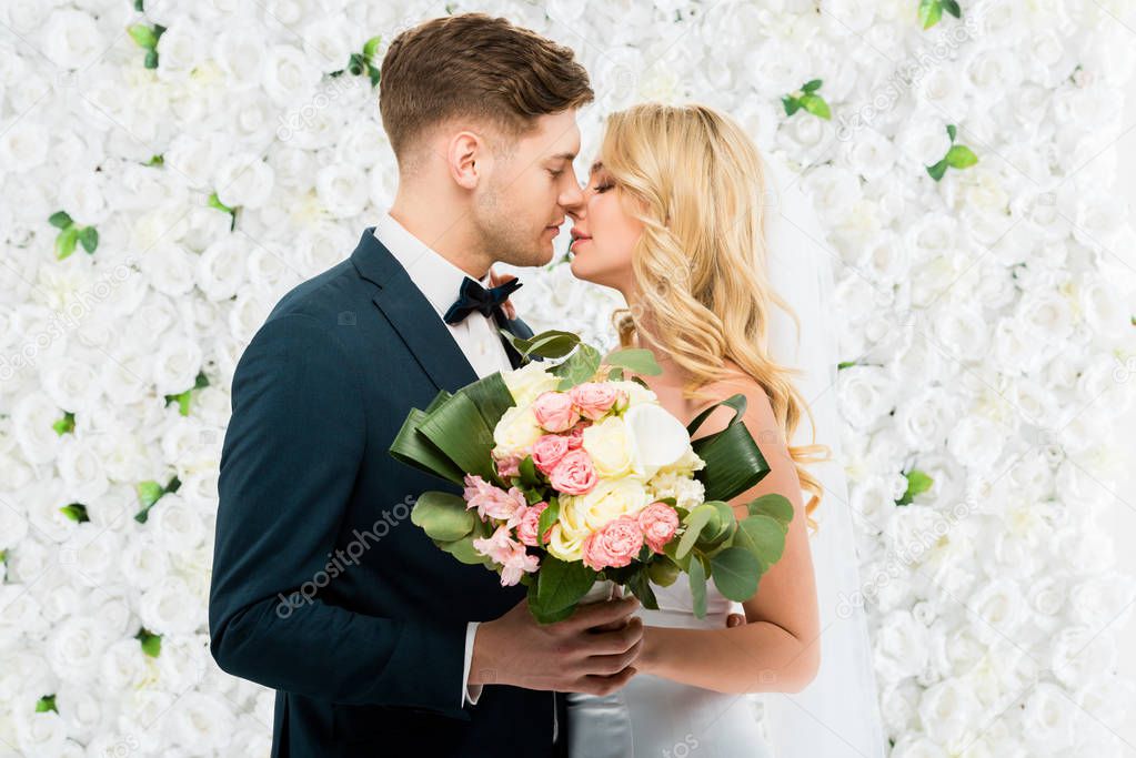 happy young bride and groom kissing while holding wedding bouquet on white floral background