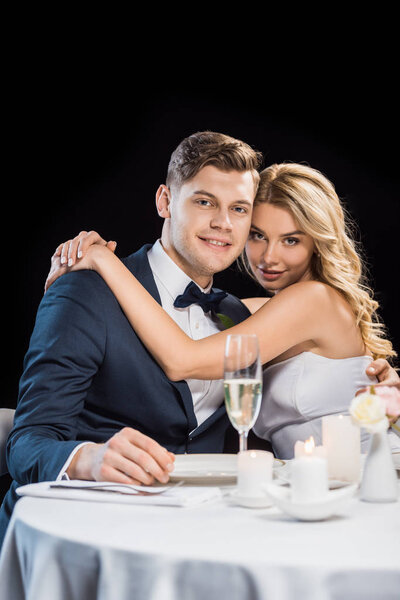 happy young groom and bride embracing and looking at camera isolated on black