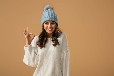 Glad smiling girl in knitted hat showing okay sign isolated on beige clipart