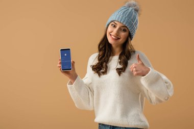 Çekici kadın smartphone ile facebook app ekran üzerinde tutarak ve izole üzerinde bej yukarı gösterilen örme şapka
