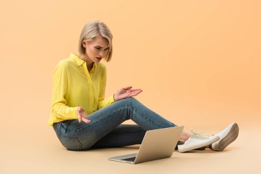 Irritated blonde girl in yellow shirt looking at laptop while sitting on orange background clipart