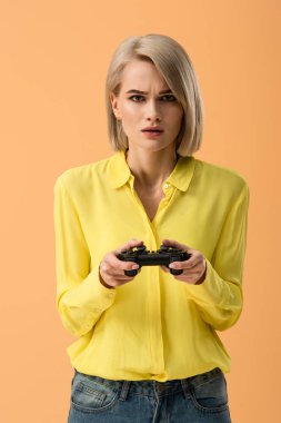 Gamepad üzerinde turuncu izole tutan sarı gömlekli endişeli sarışın kadın