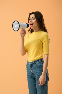 Joyful brunette girl in jeans screaming in megaphone isolated on orange clipart