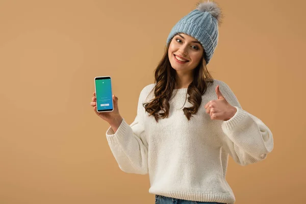 ニット帽子画面に Twitter アプリとスマート フォンを押し上孤立したベージュを親指を示す幸せなカーリー ガール — ストック写真