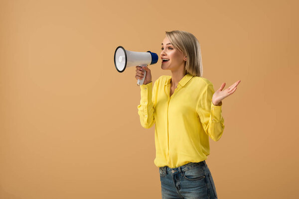 Smiling blonde woman in yellow shirt screaming in megaphone isolated on beige