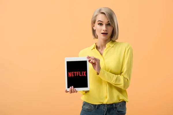 オレンジに分離された画面に Netflix アプリでデジタル タブレットを保持している黄色のシャツに驚いてのスタイリッシュな女性 — ストック写真