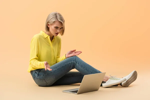 オレンジ色の背景に座ってノート パソコンを見ている黄色のシャツでイライラのブロンドの女の子 — ストック写真