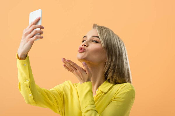 Милая блондинка посылает воздушный поцелуй, делая селфи изолированные на оранжевый
