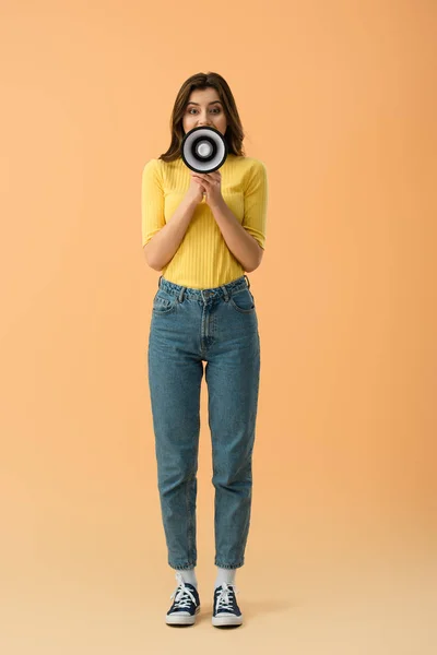オレンジ色の背景にメガホンで叫んでジーンズのブルネットの少女の完全な長さのビュー — ストック写真