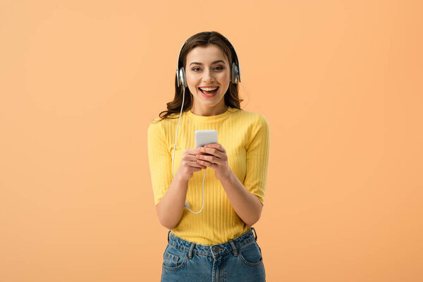 Laughing brunette girl in headphones using smartphone isolated on orange