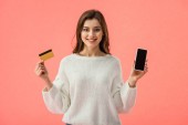 Happy brunette dívka drží kreditní kartu při pohledu na smartphone s prázdnou obrazovkou izolované na růžové 