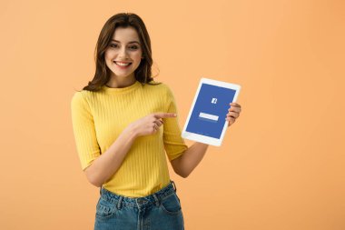 Dijital tablet ile facebook app üzerinde turuncu izole ekranda parmak ile işaret eden güzel gülümseyen kız