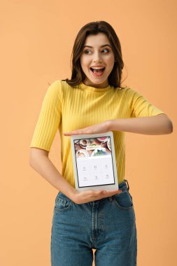 Turuncu üzerinde izole ekranda dijital tablet oturaklı app ile tutan mutlu esmer kadın