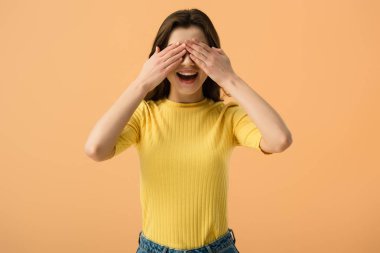 cheerful young woman covering eyes with hands isolated on orange  clipart