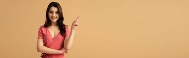 panoramic shot of cheerful brunette woman pointing with finger while standing isolated on brown  clipart