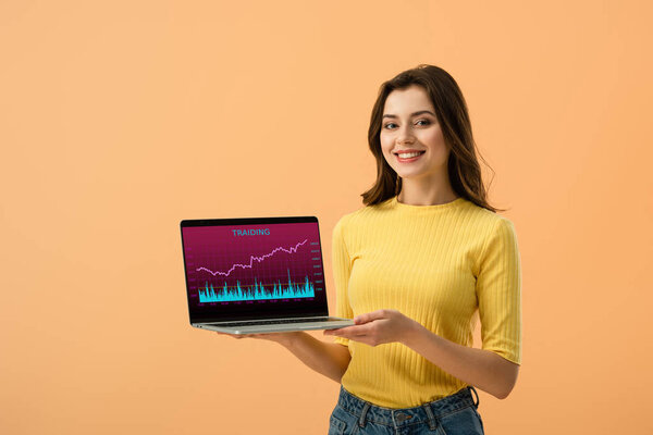 Cheerful brunette girl holding laptop with traiding website on screen isolated on orange