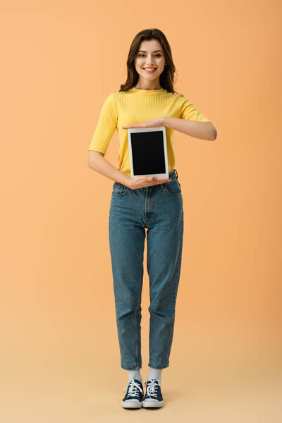 Full length view of smiling girl in jeans holding digital tablet with blank screen on orange background