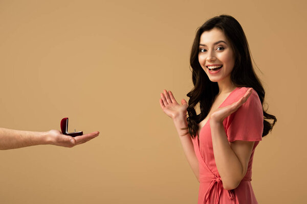 Cropped view of man holding ring and making marriage proposal to excited brunette girl isolated on beige