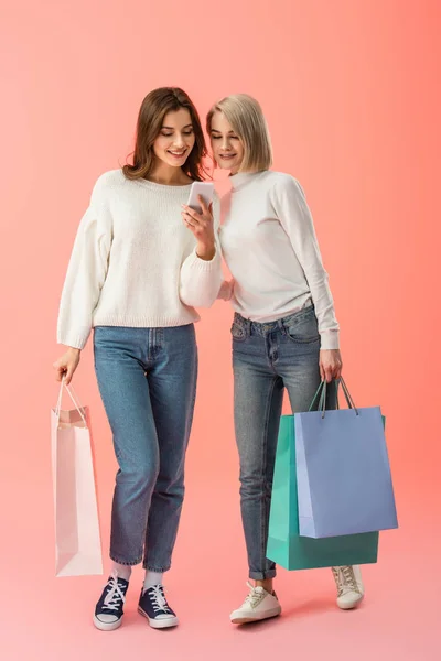 Wesoła Blondynka Brunetka Przyjaciele Patrząc Smartphone Trzymając Torby Zakupy Różowym — Zdjęcie stockowe