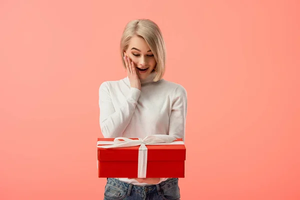 震惊的金发碧眼的年轻女子看着红色的礼品盒孤立在粉红色 — 图库照片
