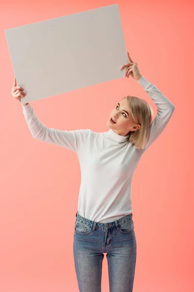 ピンクに分離された空白のプラカードを持って驚く金髪の若い女性 — ストック写真