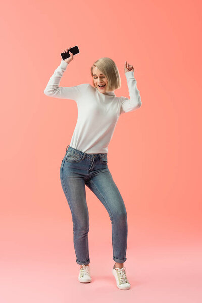 cheerful blonde girl holding smartphone and celebrating triumph on pink background