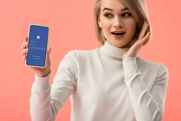 有选择的焦点惊讶的金发女子拿着智能手机与脸书应用程序在屏幕上隔离粉红色 — 图库照片