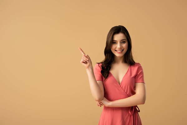 cheerful brunette woman pointing with finger while standing isolated on brown 