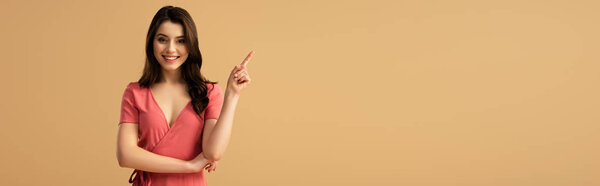 panoramic shot of cheerful brunette woman pointing with finger while standing isolated on brown 