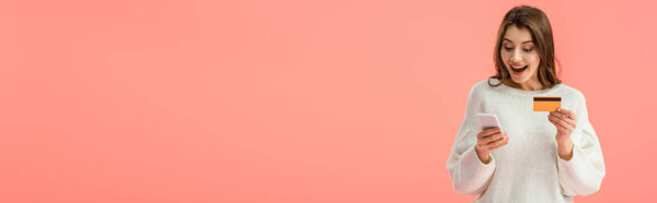 panoramic shot of surprised brunette girl holding credit card and smartphone isolated on pink