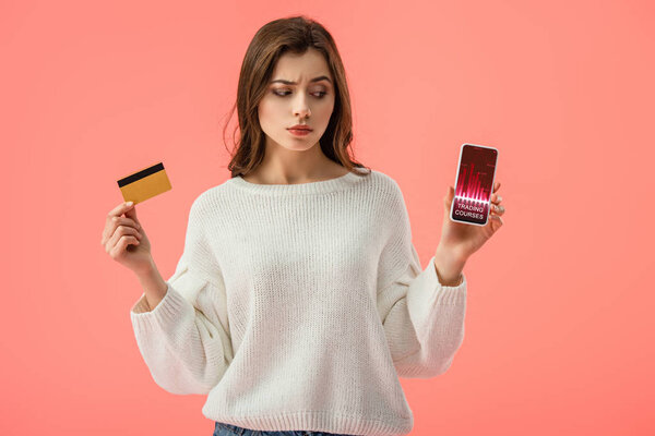 attractive brunette girl holding credit card while looking at smartphone with graphs on screen isolated on pink 