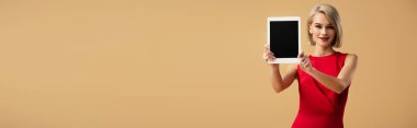panoramic shot of woman in red dress holding digital tablet with blank screen isolated on beige clipart
