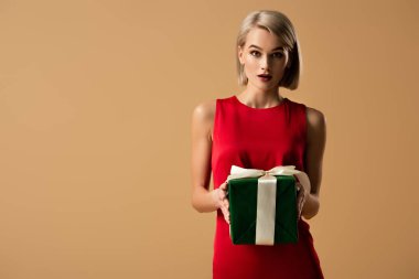 surprised and beautiful young woman in red dress holding gift box isolated on beige clipart