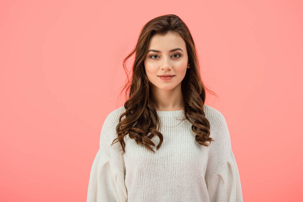 smiling and attractive woman in white sweater looking at camera isolated on pink