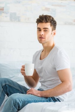 Beyaz t-shirt ve jeans yatağa oturma ve yatak odasında Kupası tutan adam