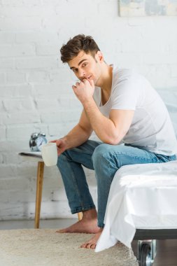 Beyaz t-shirt ve jeans yatağa oturma ve yatak odasında Kupası tutan adam