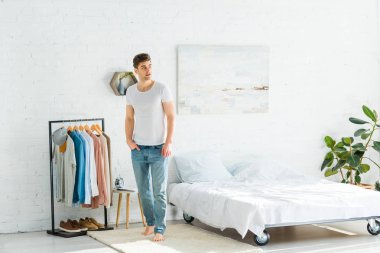 Beyaz t-shirt ve jeans yatak odasında yatak ve giysi raf yakınındaki ayakta yakışıklı adam