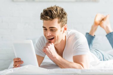 Beyaz t-shirt dijital tablet tutarak ve yatak odasında gülümseyen yatakta yakışıklı adam