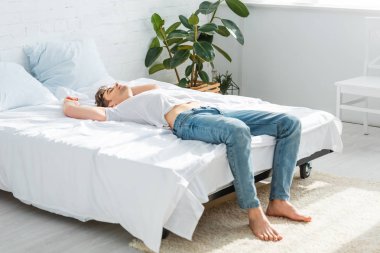 handsome man in t-shirt and jeans lying on back on bed in bedroom  clipart