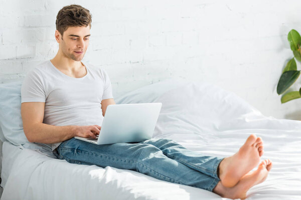 красивый мужчина в белой футболке и джинсах сидит на кровати и печатает на ноутбуке в спальне
