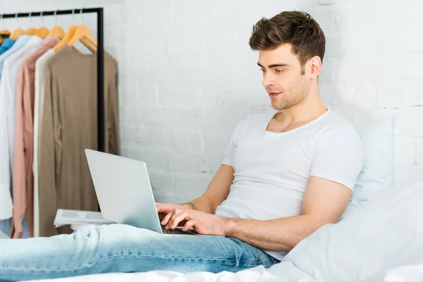 Hombre Camiseta Blanca Jeans Sentado Cama Escribiendo Ordenador Portátil Dormitorio — Foto de Stock