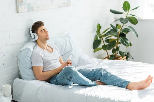 Handsome Man White Shirt Jeans Sitting Bed Holding Smartphone Listening — Stock Photo, Image