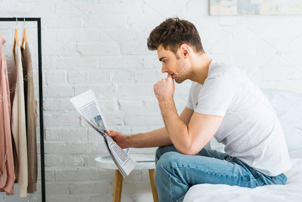 man in white t-shirt and jeans sitting on bed and reading newspaper at home