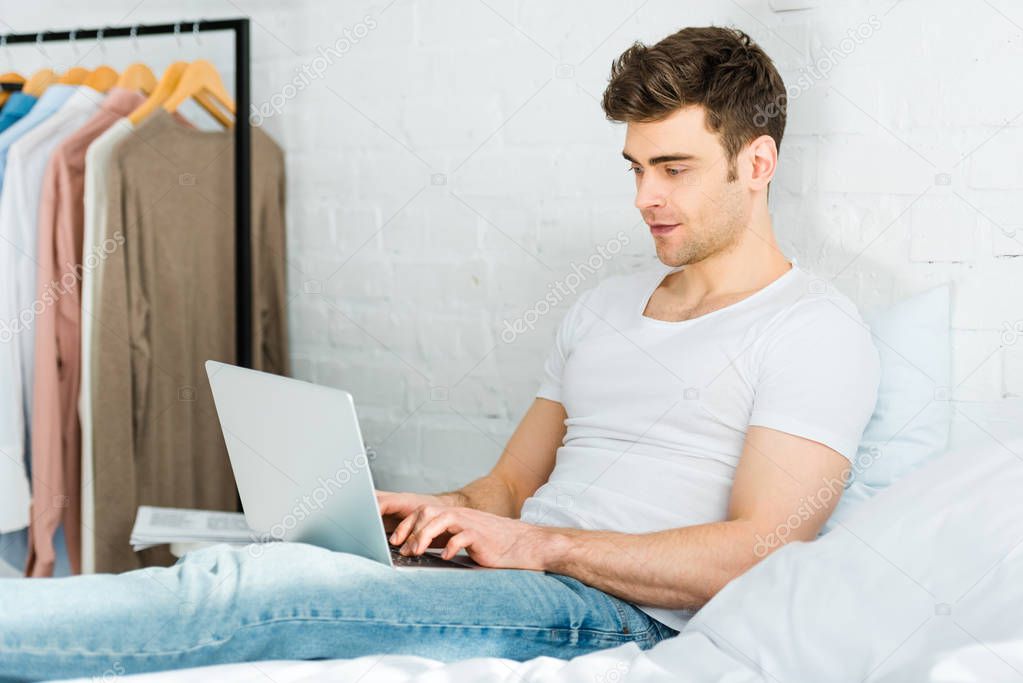 man in white t-shirt and jeans sitting on bed and typing on laptop in bedroom