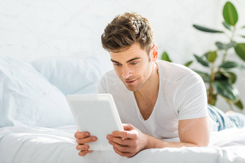 handsome man in white t-shirt lying on bed with digital tablet in bedroom