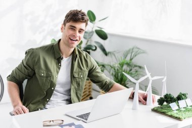 çimenlerin üzerinde laptop, yel değirmenleri, güneş panelleri modelleri ile masada oturan ve ofiste gülümseyen yakışıklı mimar