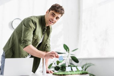 handsome architect standing and putting solar panel model on grass near trees, windmills model and laptop on table in office clipart