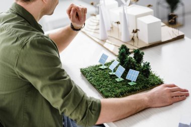 cropped view of architect sitting at table with solar panels and trees models in office clipart