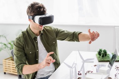 architect in virtual reality headset sitting at table with laptop, windmills and trees models in office clipart