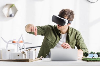Tablo ile laptop ve yel değirmenleri, binalar, ağaçlar, güneş panelleri modelleri ofiste oturup siyah sanal gerçeklik kulaklık mimar